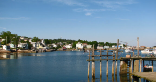 A serene coastal scene with a calm harbor, wooden docks, and charming houses lining the shore under a clear blue sky.