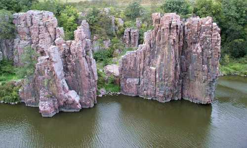 Rock formations rise above a calm body of water, surrounded by greenery and trees in a natural landscape.