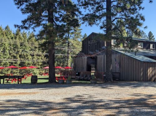 A rustic wooden building surrounded by tall trees, with outdoor seating and red umbrellas in a sunny setting.