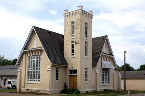 A historic yellow brick church with a tall tower and large windows, set against a cloudy sky.