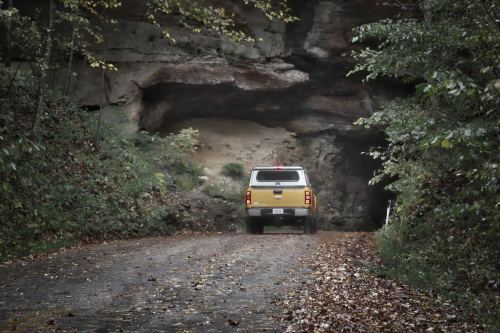 A yellow truck drives on a gravel road leading into a rocky cave surrounded by trees and fallen leaves.