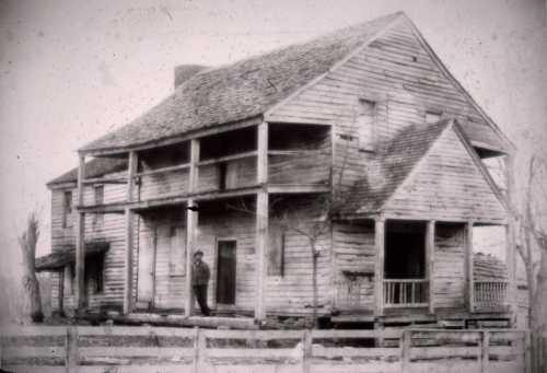 A historic wooden house with a porch, featuring a man standing at the entrance, surrounded by a fence and trees.