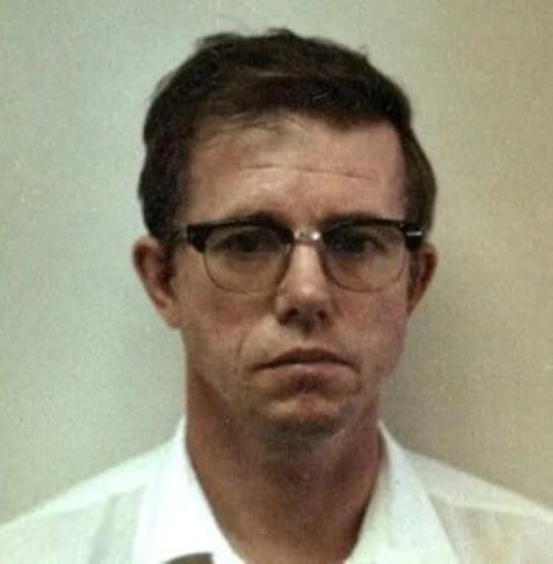 A serious-looking man with glasses and short hair, wearing a white shirt, against a plain background.