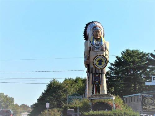 A large statue of a Native American chief stands beside a road, surrounded by trees and signage.
