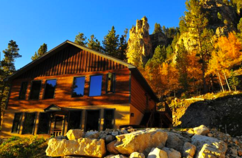 A modern wooden cabin surrounded by autumn trees and rocky cliffs under a clear blue sky.