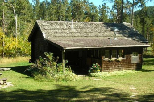 A rustic wooden cabin surrounded by trees and greenery, with a clear blue sky above.