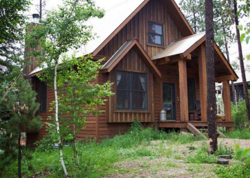 A rustic wooden cabin surrounded by trees and greenery, featuring a porch and large windows.