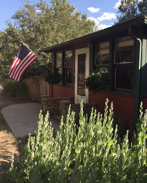 A cozy red house with a porch, featuring an American flag and potted plants, surrounded by greenery.