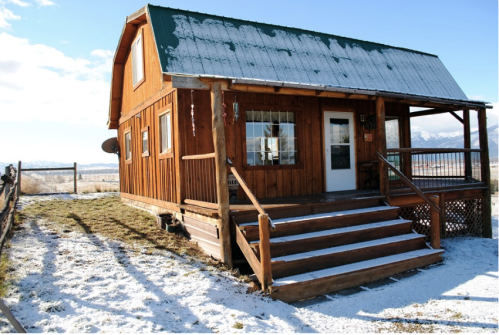 A rustic wooden cabin with a snow-covered roof, surrounded by a snowy landscape and mountains in the background.