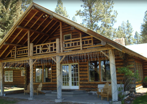 A rustic log cabin with a large porch, surrounded by trees and natural scenery.