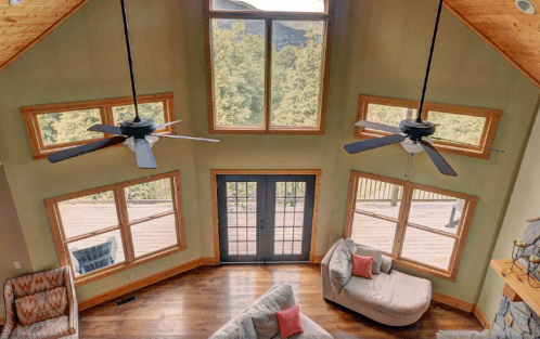 Spacious living room with large windows, two ceiling fans, and cozy seating, overlooking a deck and greenery outside.