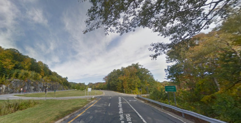 A scenic road curves through a forested area with autumn foliage under a partly cloudy sky.