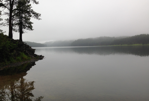 A calm lake surrounded by misty hills and trees, reflecting the serene landscape on the water's surface.