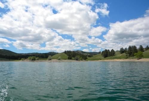 A serene lake surrounded by green hills and trees under a partly cloudy blue sky.