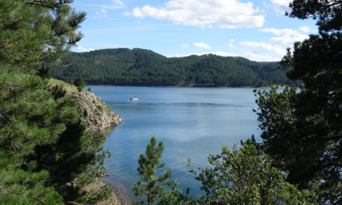 A serene lake surrounded by green trees and mountains, with a small boat on the water under a blue sky.