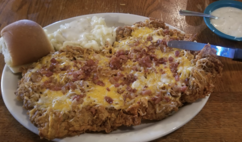 A plate featuring a large fried chicken dish topped with cheese and bacon, served with mashed potatoes and a roll.