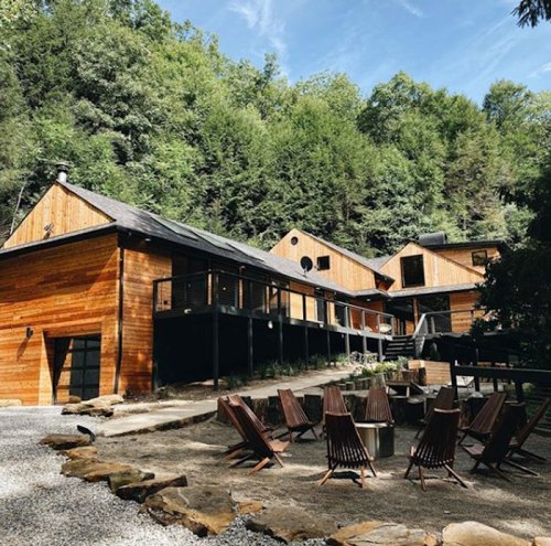 A modern wooden cabin surrounded by trees, featuring a patio with chairs and a gravel pathway.