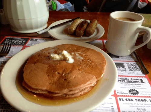 A plate of pancakes with butter and syrup, accompanied by sausages and a cup of coffee on a table with menus.
