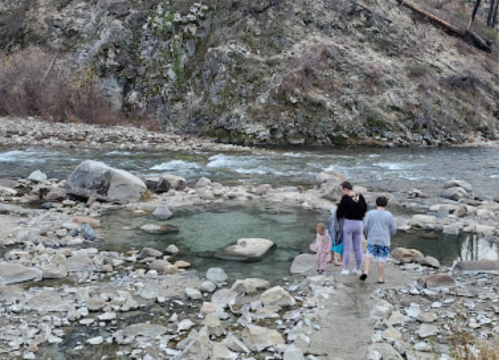 A family walks along a rocky riverbank, with a backdrop of a hillside showing signs of fire damage.