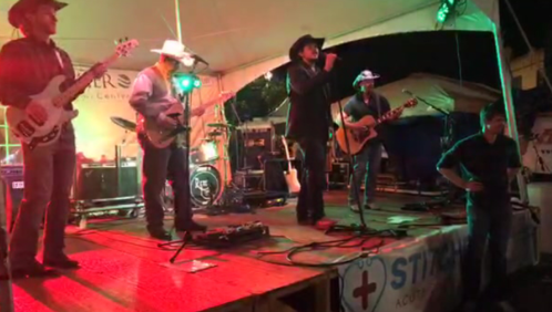 A country band performs on stage under a tent, with musicians playing guitar and bass, and a lively audience.