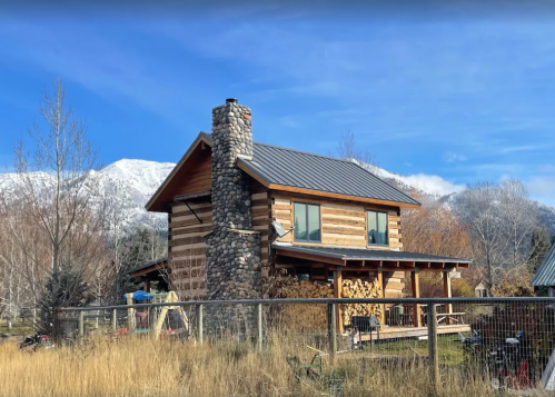 A rustic log cabin with a stone chimney, surrounded by grass and mountains under a clear blue sky.