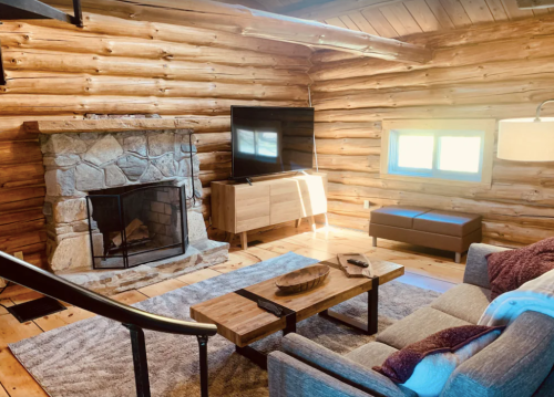 Cozy log cabin interior with a stone fireplace, TV, wooden furniture, and natural light streaming through the windows.
