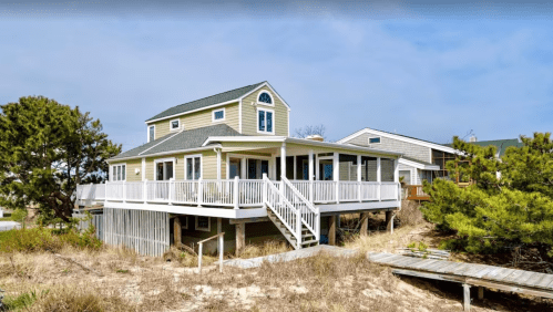 A coastal house with a green exterior, elevated on stilts, surrounded by sand and greenery.
