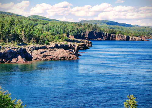 A serene coastal landscape featuring rocky cliffs, lush greenery, and calm blue waters under a partly cloudy sky.