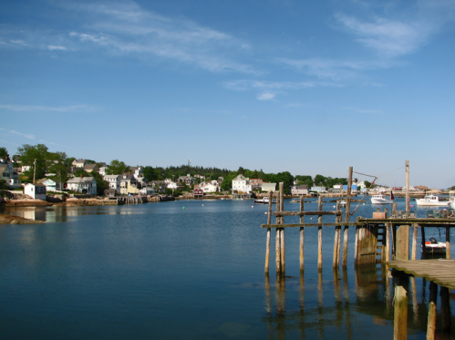 A serene coastal scene with a calm blue harbor, wooden docks, and quaint houses lining the shore under a clear sky.