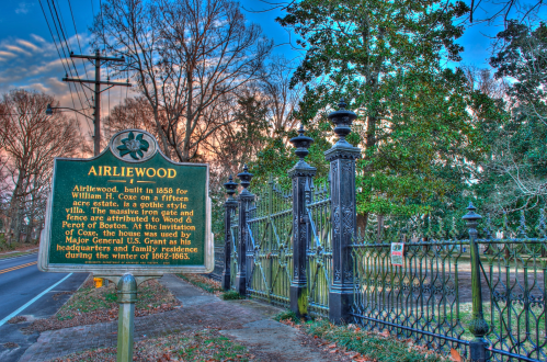 Historic sign for Airliewood, a Gothic-style estate built in 1855, with a wrought iron gate and trees in the background.