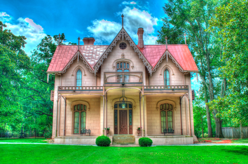 A charming Victorian-style house with a red roof, intricate details, and surrounded by lush greenery under a blue sky.