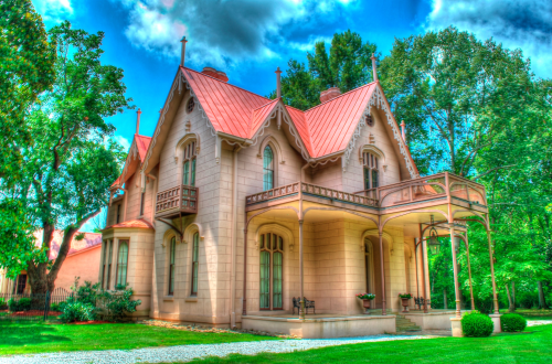 A charming Victorian-style house with a red roof, surrounded by lush greenery and a clear blue sky.