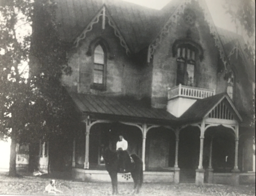 A historic black-and-white photo of a man on a horse in front of a large, ornate house with a dog nearby.