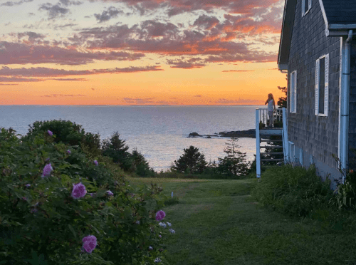 A woman stands on a porch overlooking a serene ocean at sunset, with colorful clouds and lush greenery in the foreground.