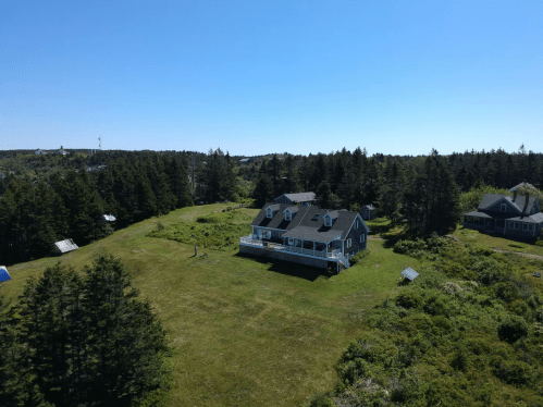 Aerial view of a large house on a grassy hill surrounded by trees under a clear blue sky.