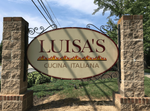 Sign for "Luisa's Cucina Italiana" featuring decorative flames and a stone base, set against a green landscape.