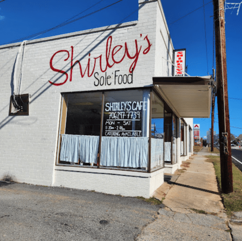 Exterior of Shirley's Sole Food restaurant, featuring a sign and large windows with curtains, on a sunny day.