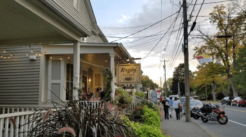 A quaint tavern with a sign, surrounded by greenery, people walking by, and a cloudy sky in the background.