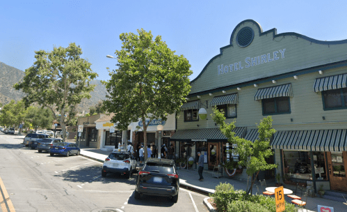 A street view of Hotel Shirley and nearby shops, with people gathered outside and trees lining the sidewalk.