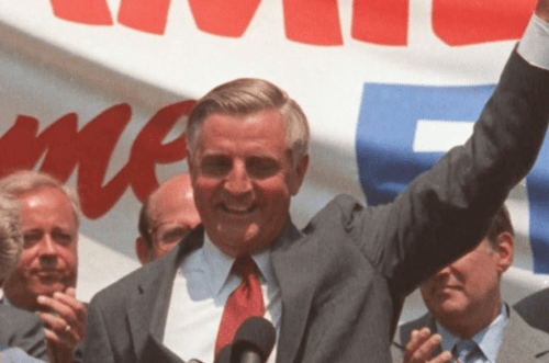 A smiling man in a suit raises his hand in a celebratory gesture at a public event, with supporters in the background.