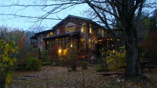 A cozy wooden house surrounded by trees and autumn foliage, illuminated softly at dusk.