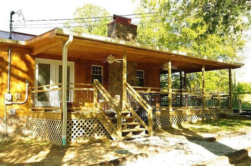 A wooden house with a spacious porch, stone accents, and steps leading up, surrounded by trees and greenery.