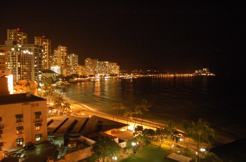 Nighttime view of a beachside city with illuminated buildings and calm water reflecting city lights.