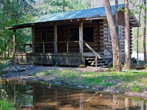 A rustic log cabin surrounded by trees, with a small creek running nearby and a porch in front.