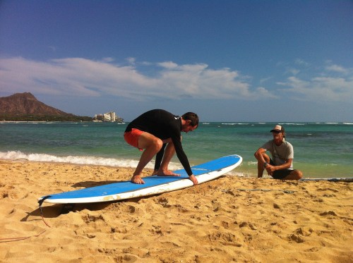 A man prepares to surf on a blue board while another man sits on the beach, with ocean and mountains in the background.