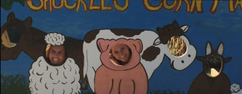 Two smiling people pose through cutouts of a sheep and a pig, with painted cows and goats in the background.
