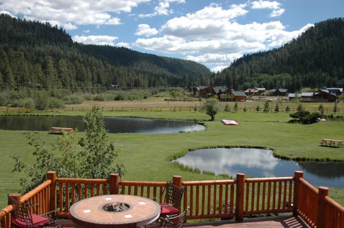 Scenic view of a lush green landscape with ponds, mountains, and a wooden deck in the foreground under a blue sky.