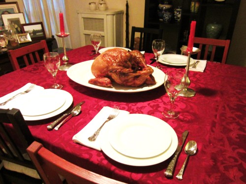 A festive dining table set with plates, cutlery, and a large roasted turkey centerpiece. Red candles add ambiance.