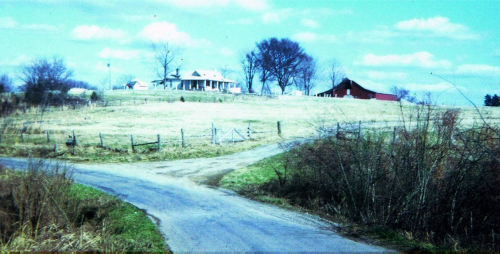 A rural scene featuring a house and barn on a grassy hill, with a winding road and trees under a blue sky.