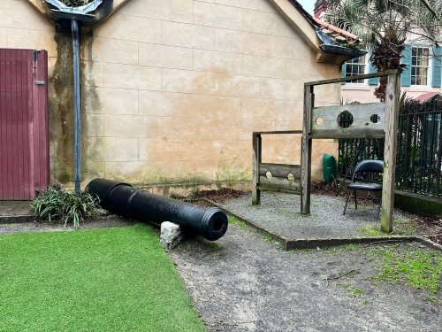 A black cannon lies on the ground next to a wooden structure with holes, beside a chair and a grassy area.